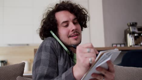 Close-up-focused-happy-brunette-guy-with-curly-hair-and-mustache-in-a-checkered-shirt-listens-communicates-on-the-phone-and-writes-down-important-points-in-a-notepad-during-his-working-day-at-home