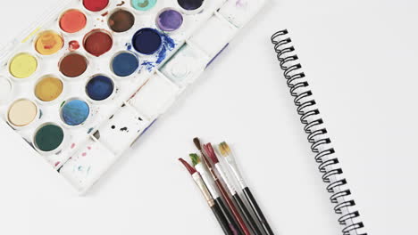 primer plano de un cuaderno, pinturas y pinceles sobre un fondo blanco, en cámara lenta