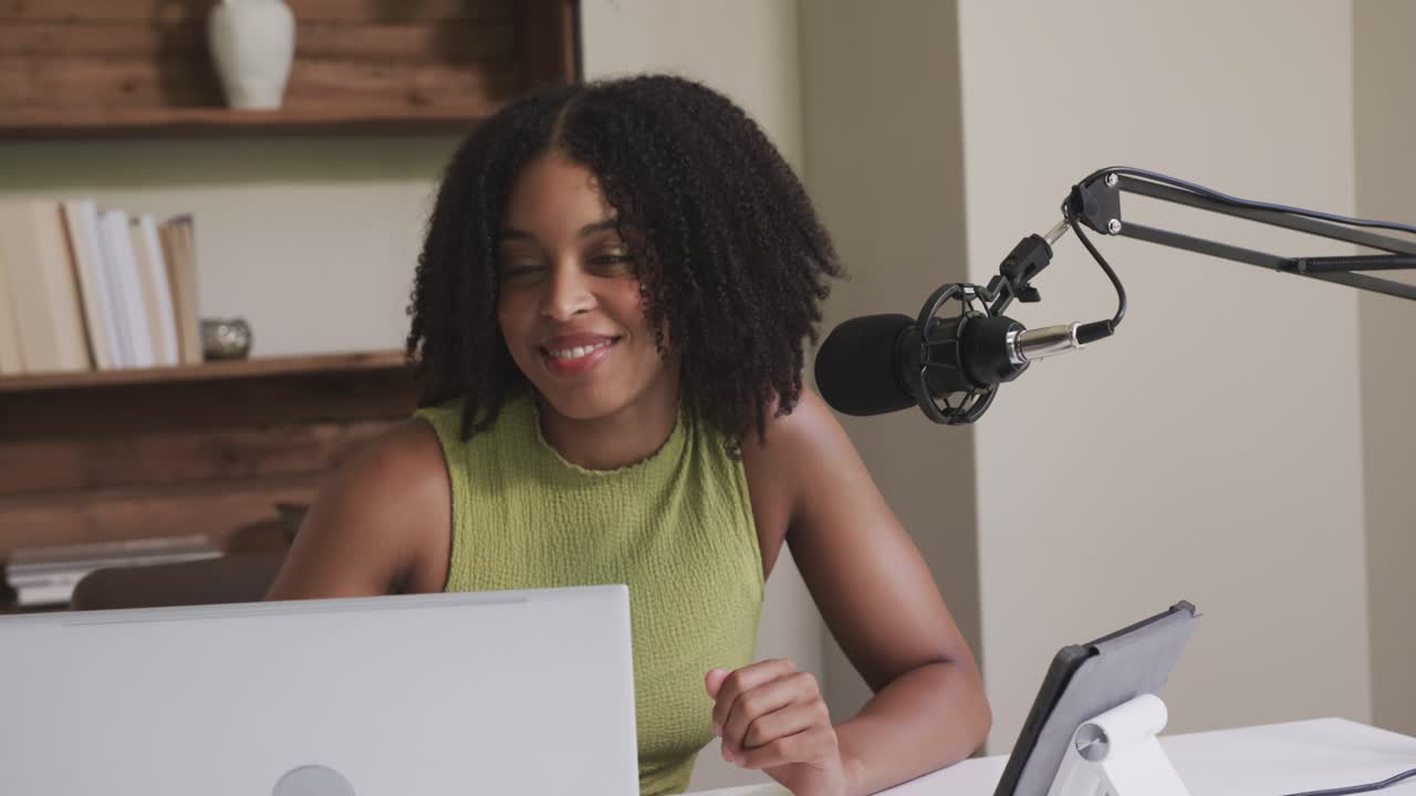 African American woman taking breath, recording into mic and consulting laptop at desk in green top