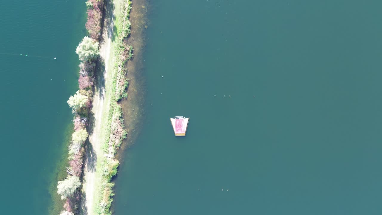 Aerial downward view of a dike dividing the lake. Trees line a dirt road on the dike. A water ski slalom course with ramp and buoys is clearly visible