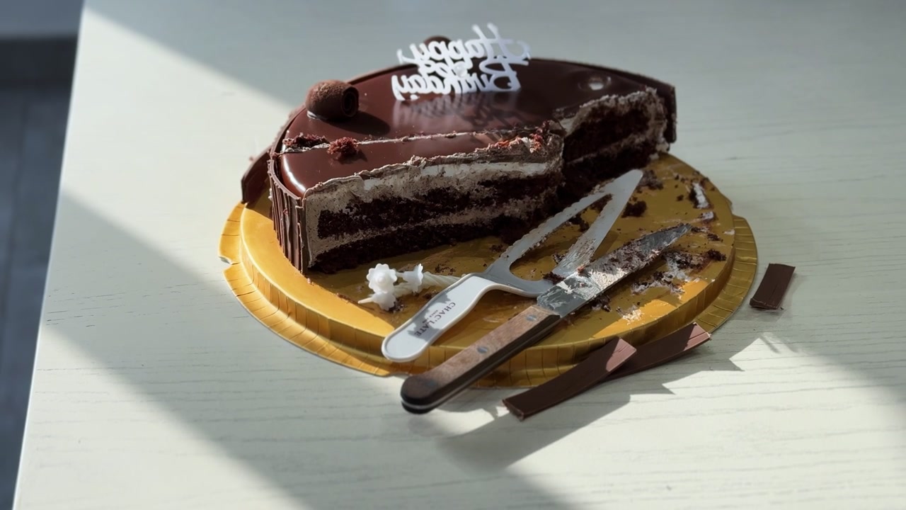 Un primo piano di una torta di compleanno al cioccolato a fette su un tavolo bianco da interni. Il dessert mezzo mangiato è illuminato dalla luce del giorno accanto a una paletta per dolci, un coltello e briciole di cacao sparse