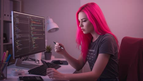 a young female programmer with vibrant pink hair engaged in coding, focused on her computer screen while holding a pen in a modern workspace setting