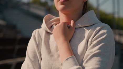 close-up of woman s hand gently holding hoodie, with sunlight casting a warm glow on her face, the background is blurred, featuring an empty stadium and trees