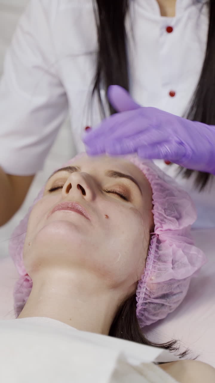 An experienced cosmetologist in sterile gloves makes a face massage to a woman on the couch in the cosmetology office. Relax. Facial Moisturizing Procedure Vertical video