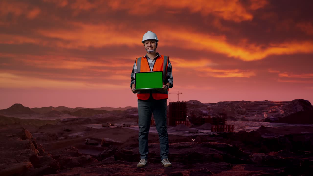 cuerpo lleno de ingeniero masculino asiático con casco de seguridad sonriendo y mostrando portátil de pantalla verde a la cámara mientras estaba de pie en el sitio de construcción, maquinaria de minería
