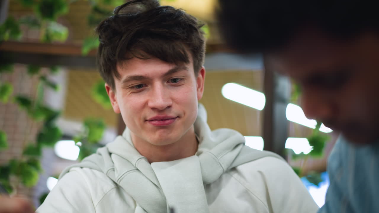 Teenager adjusting white sweater around neck while admiring bestie sketching detailed drawing on sketchbook at wooden desk with bright vibrant warm natural soft light friendship close up art session