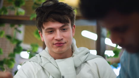 teenager adjusting white sweater around neck while admiring bestie sketching detailed drawing on sketchbook at wooden desk with bright vibrant warm natural soft light friendship close up art session