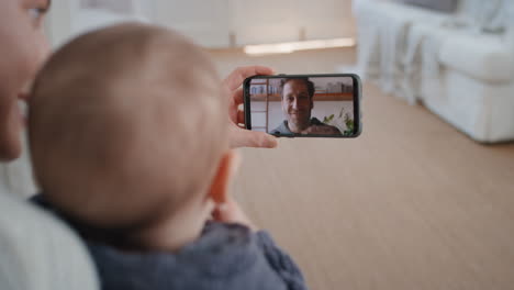 Mutter-Und-Baby-Unterhalten-Sich-Per-Smartphone-Per-Video-Chat-Mit-Dem-Vater,-Der-Dem-Säugling-Auf-Dem-Bildschirm-Zuwinkt-Und-Genießen-Die-Kommunikation-Mit-Der-Familie-über-Eine-Mobiltelefonverbindung-In-4K