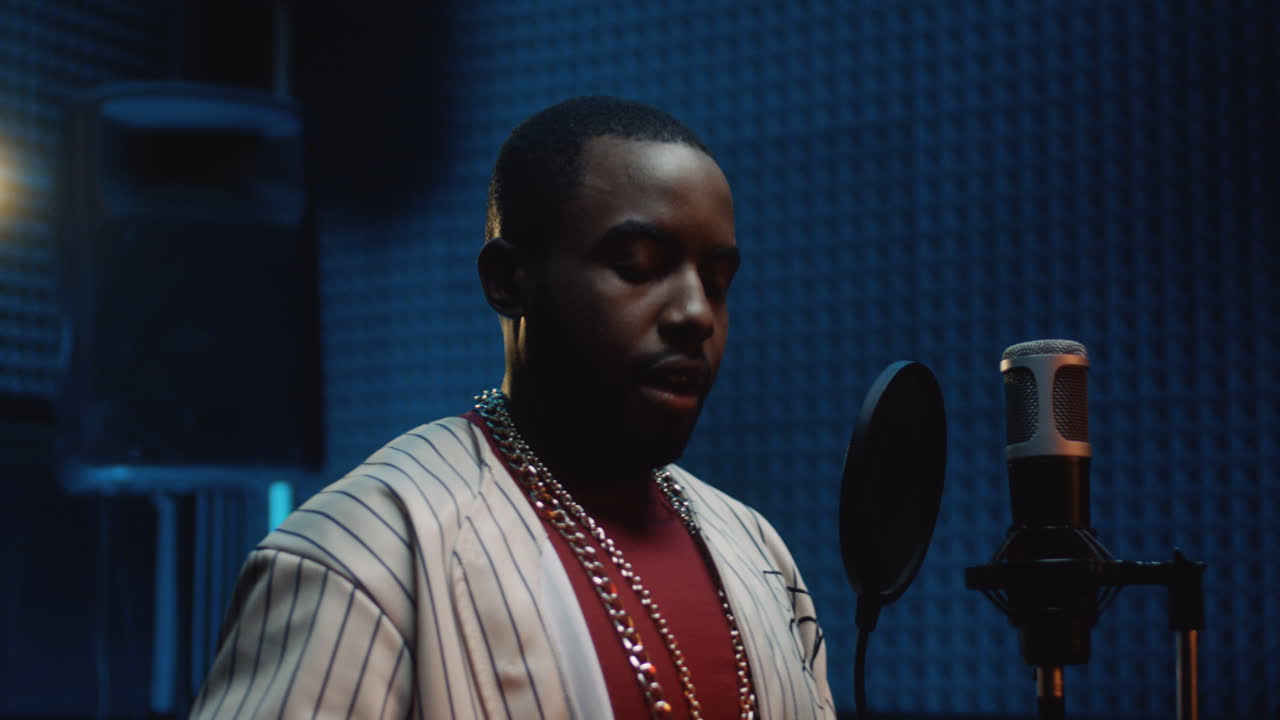 젊은 세련된 남자 랩퍼가 헤드폰을 끼고 어두운 사운드 스튜디오에서 노래를 녹음하기 시작합니다.