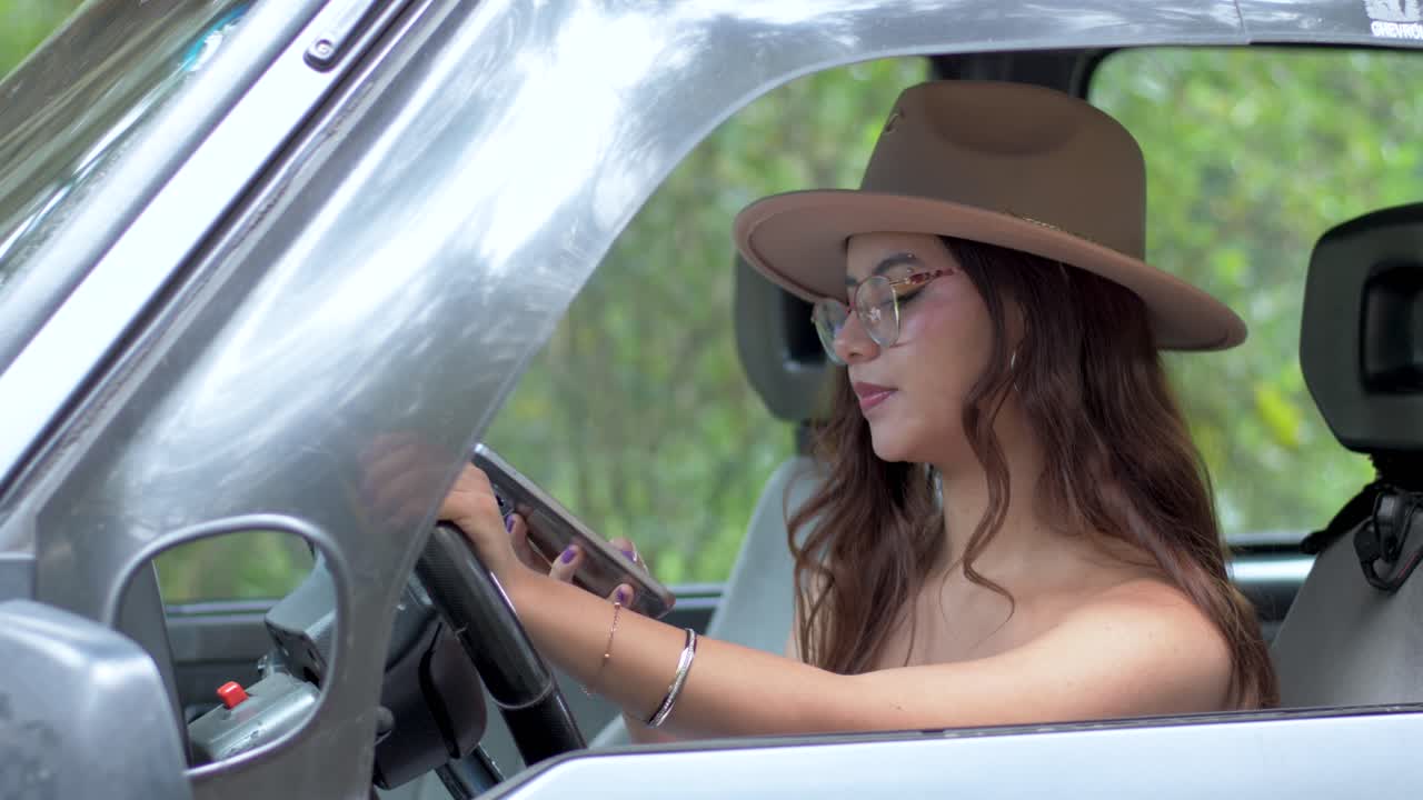 una chica atractiva está sentada detrás del volante de un coche y mirando ocupada en el teléfono y escribiendo mensajes de texto