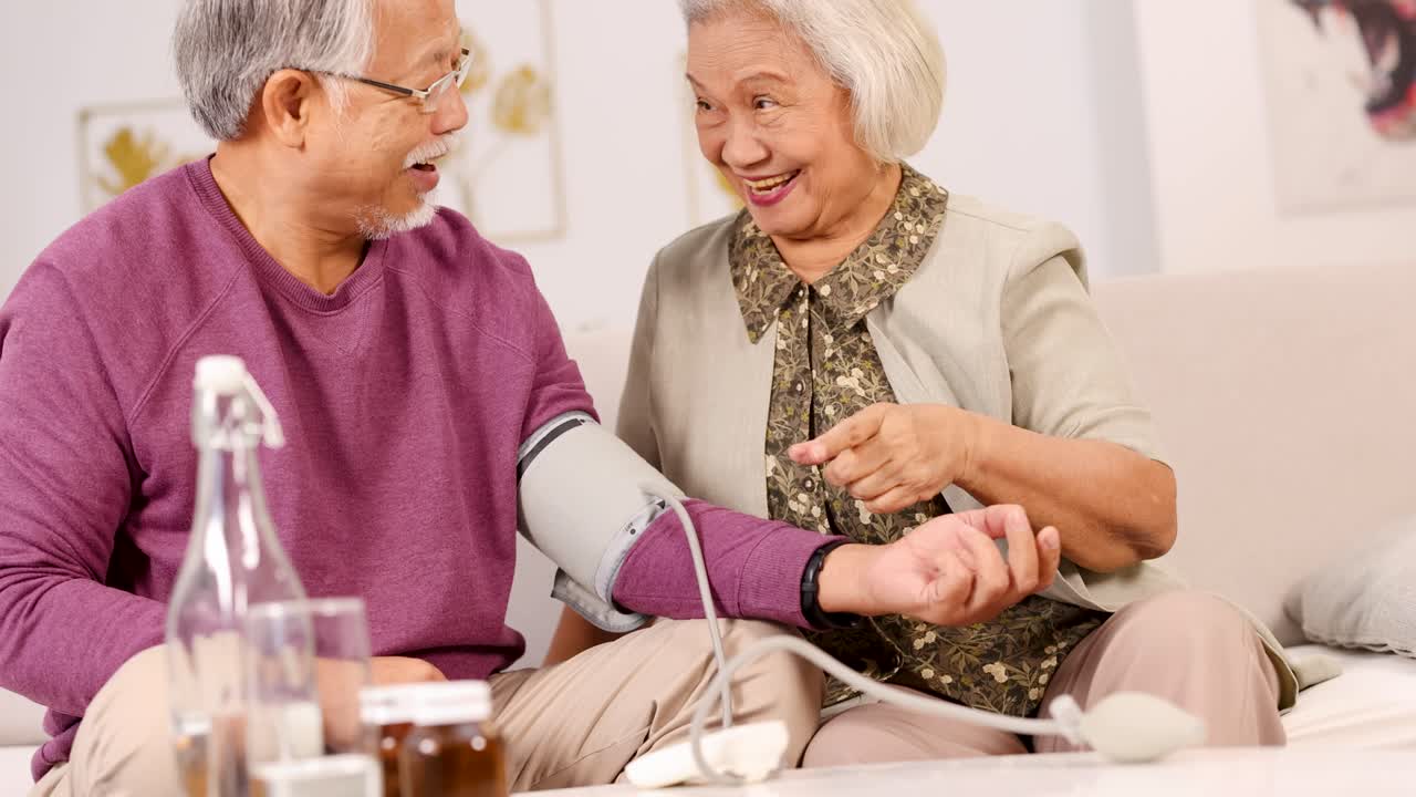 Elderly couple measuring blood pressure at home, laughing together in a bright, warm living room