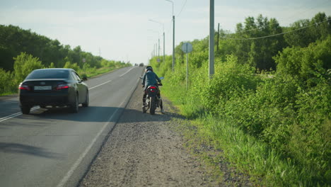 a back view of a woman climbing onto a motorcycle parked by the roadside, with lush green bushes in the background and cars approaching from behind along the road