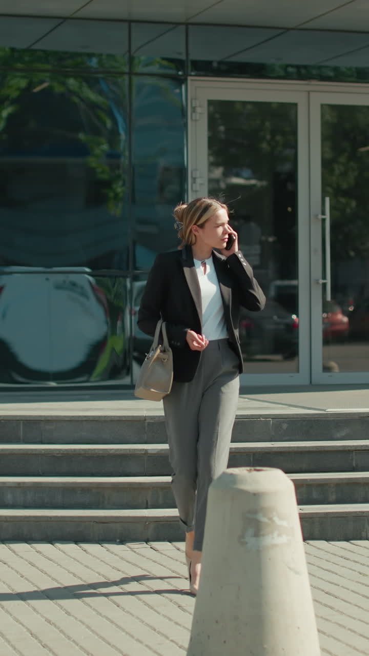 Banker on phone call stepping down staircase outside modern glass office building, holding handbag with sleek reflections of cars and surroundings on facade