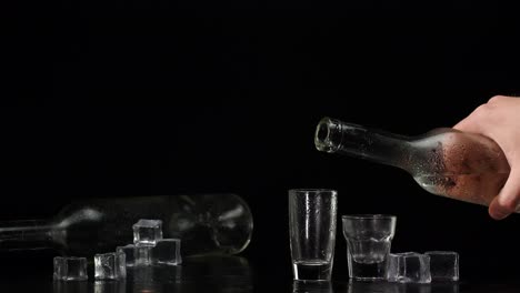 Bartender-pouring-up-two-shots-of-vodka-with-ice-cubes-from-bottle-into-glasses-on-black-background