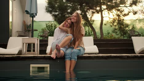 Two-girlfriends-are-sitting-near-the-sunbeds-by-the-pool,-hugging-and-looking-into-the-distance.-Rest-in-the-country-house