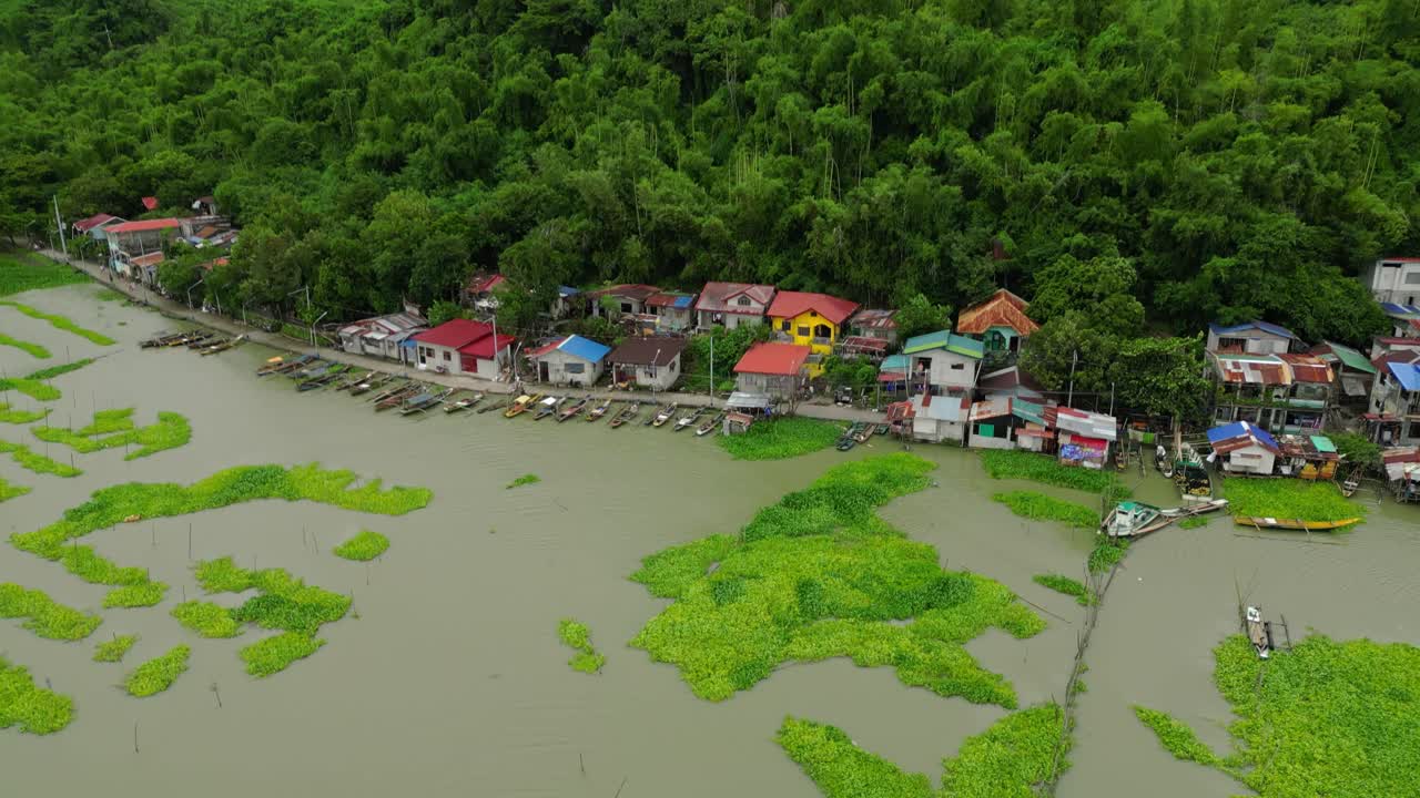 Drone shot glides over a Philippine riverside village with colorful rooftops, docked boats, and floating vegetation, framed by dense forested hills and calm waters