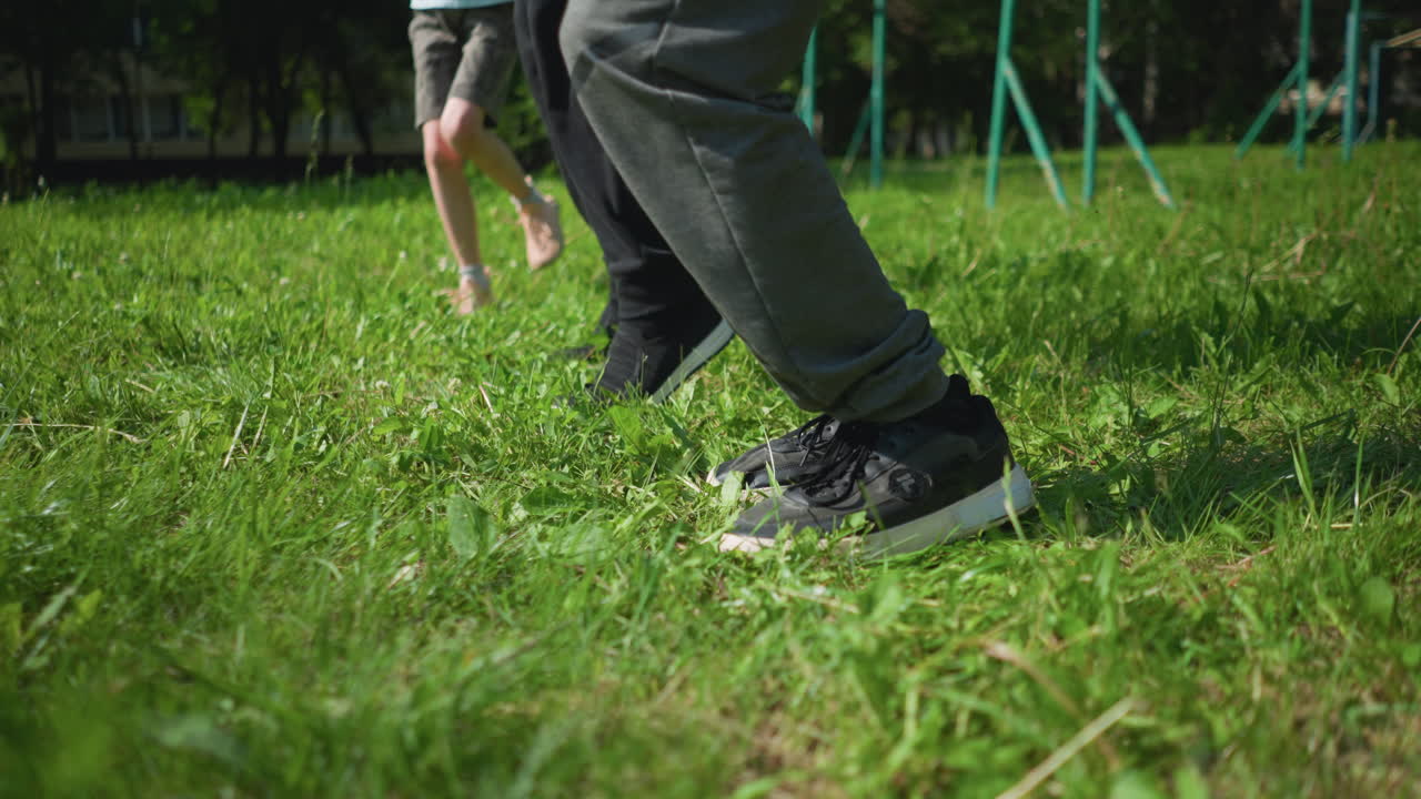 Close-up of two adults jumping during an outdoor sports exercise on a grassy field, with a young girl hopping on one leg, with a blur view of outdoor equipment behind them