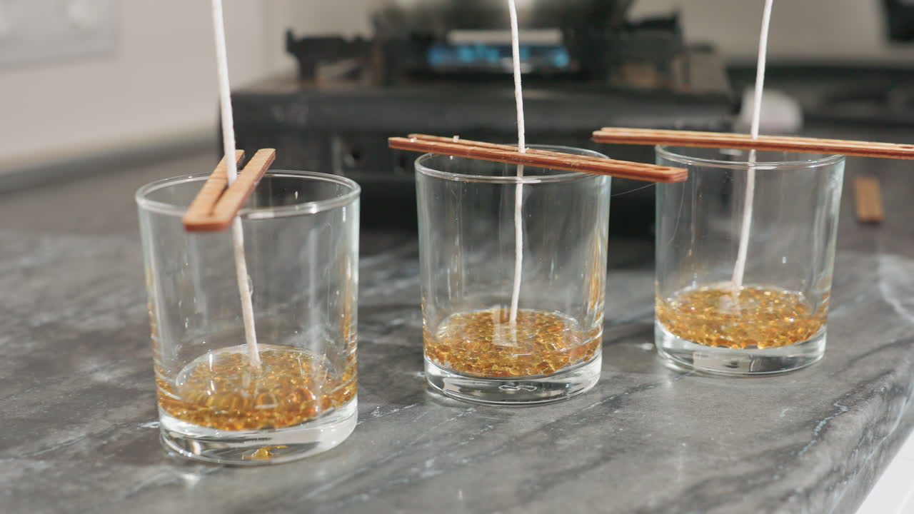 Close up of three glass cups with candle wicks held upright by wooden chopsticks resting across rims, ready for pouring wax during homemade candle production, with soft blurred background