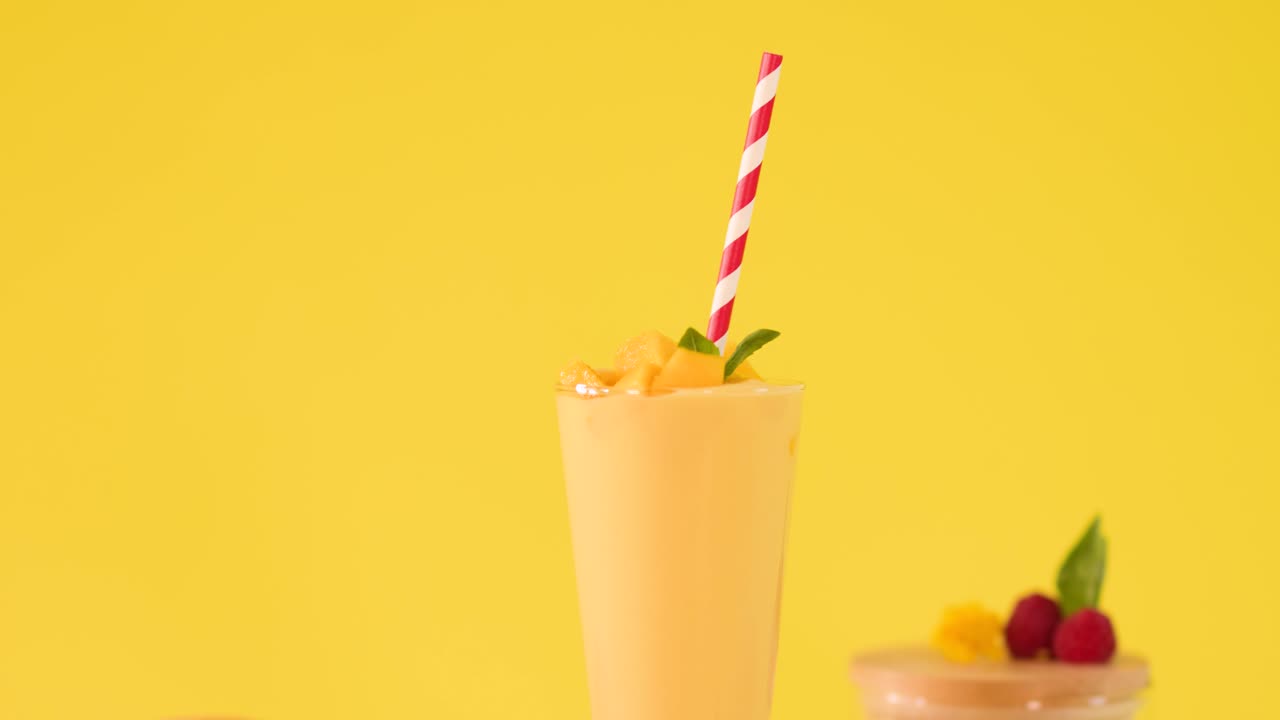 Una mano umana decora un frullato freddo al mango con un topper di carta a forma di frutta su uno sfondo giallo brillante