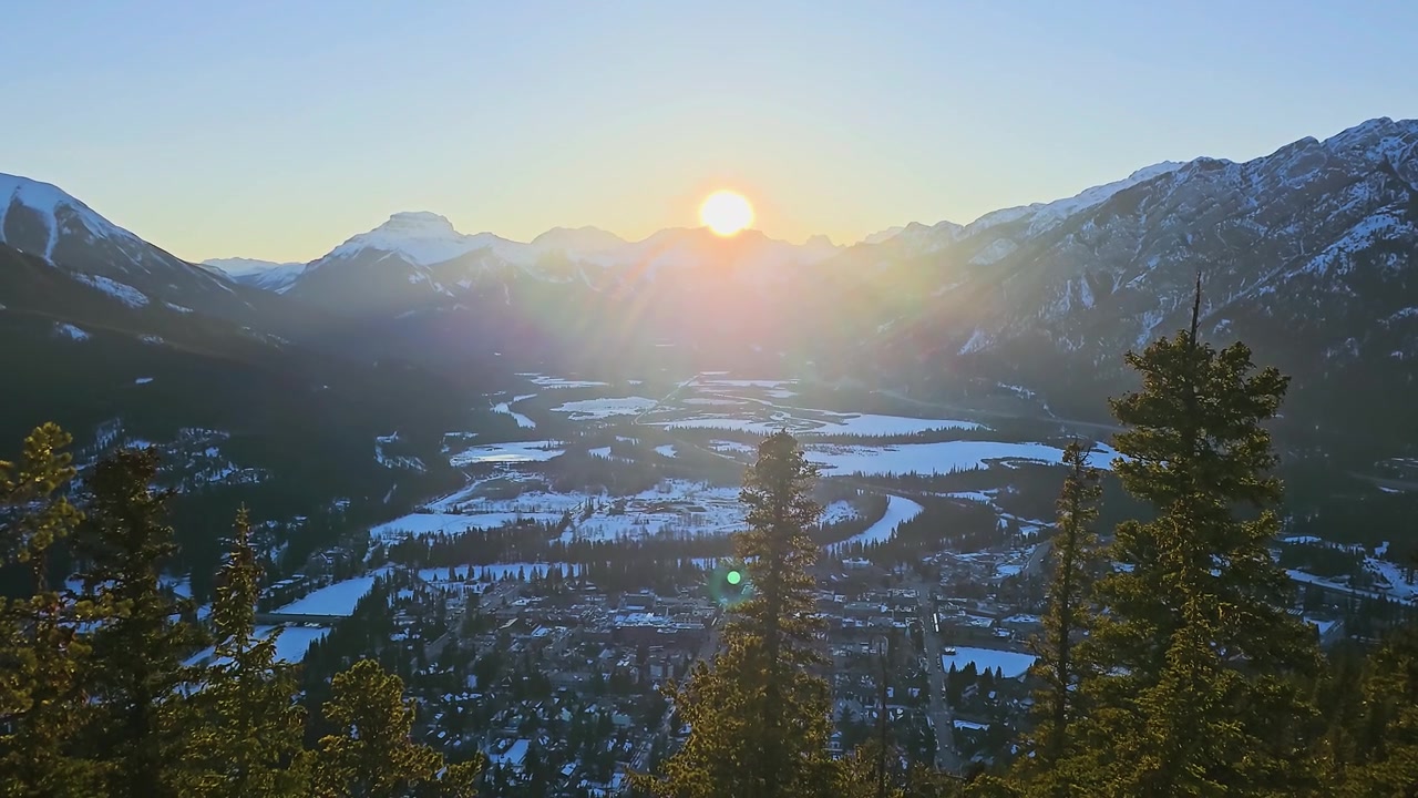 Toàn cảnh núi mùa đông tại Vườn quốc gia Banff, Canada, với mặt trời thấp rực rỡ, đỉnh núi tuyết, cây thường xanh, thung lũng đóng băng, thị trấn nghỉ dưỡng tuyệt đẹp bên dưới và du lịch Dãy núi Rocky.