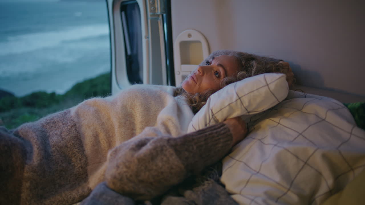 Campingkvinde ligger i seng i en campingvogn ved stormfulde havbølger i skumringen. Nærbillede.