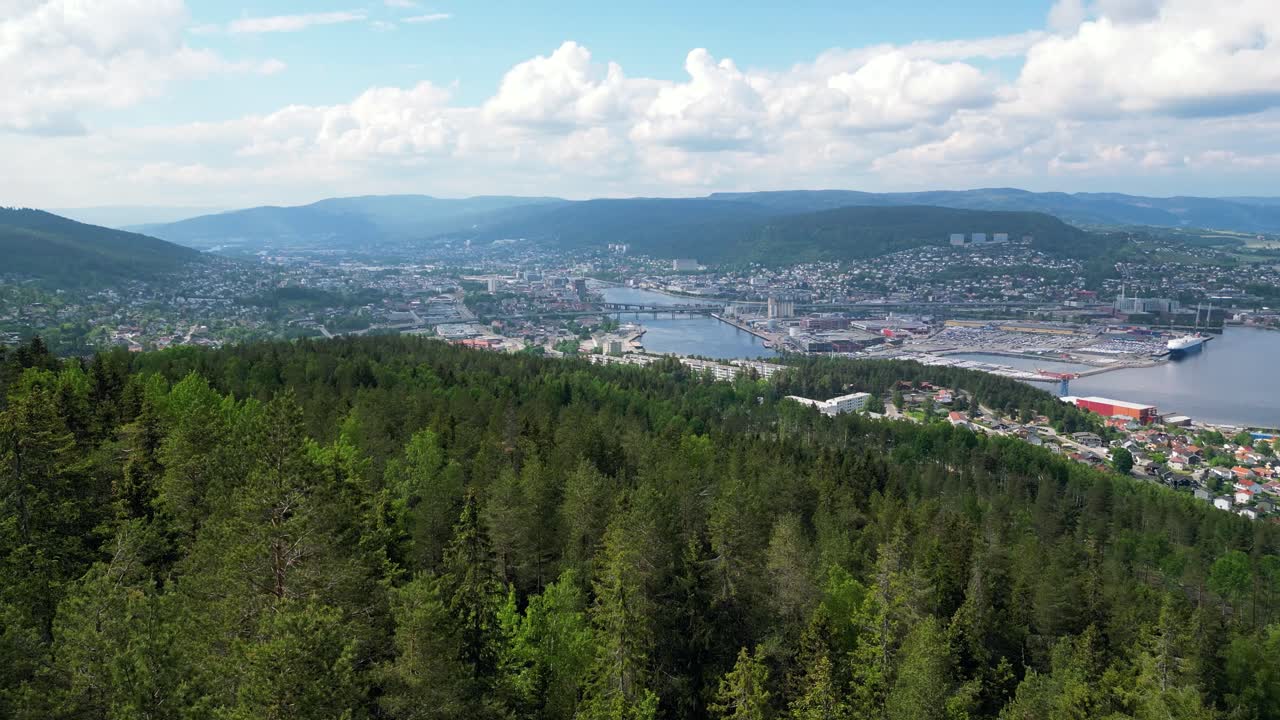 imágenes aéreas de drammen, noruega, destacando el contraste entre el denso bosque en primer plano y el vibrante paisaje urbano y el río en el fondo bajo un cielo azul brillante