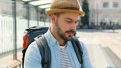 joven y apuesto viajero caucásico con sombrero con mochila sosteniendo un mapa de la ciudad y planificando una ruta en la calle