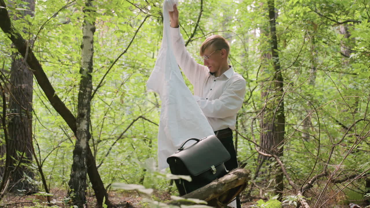 Young man in white shirt stands among green trees folding coat into bag while surrounded by dense forest vegetation, natural light filtering through branches creating tranquil outdoor atmosphere