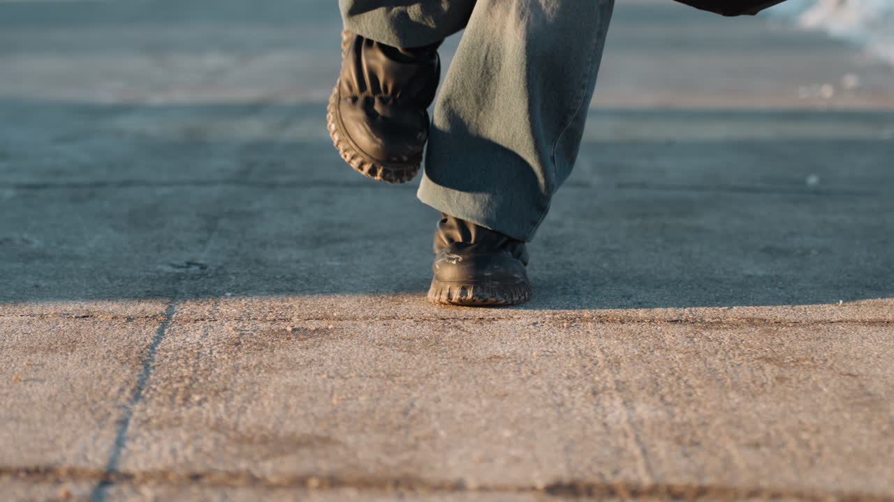 Piedi in primo piano che camminano su un pavimento crepato di un tetto, stivali consumati e denim, passi lenti e deliberati, fessure di cemento testurizzate, luce soffusa del crepuscolo, lunga ombra, umore solitario e resiliente, battistrada consumato.