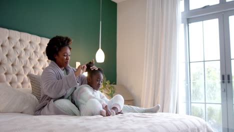 happy african american mother combing daughter hair sitting on bed in bedroom, slow motion