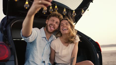 A-bearded-guy-with-curly-hair-in-a-blue-shirt-is-smiling,-grimacing-at-the-camera-with-his-blonde-girlfriend-in-a-white-top-and