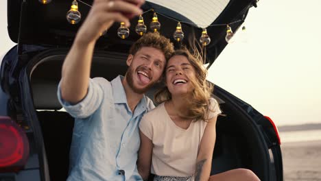 a bearded guy with curly hair in a blue shirt is smiling, grimacing at the camera with his blonde girlfriend in a white top and