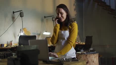 caucasian female jeweller in workshop wearing apron, sitting at desk, using laptop and smartphone