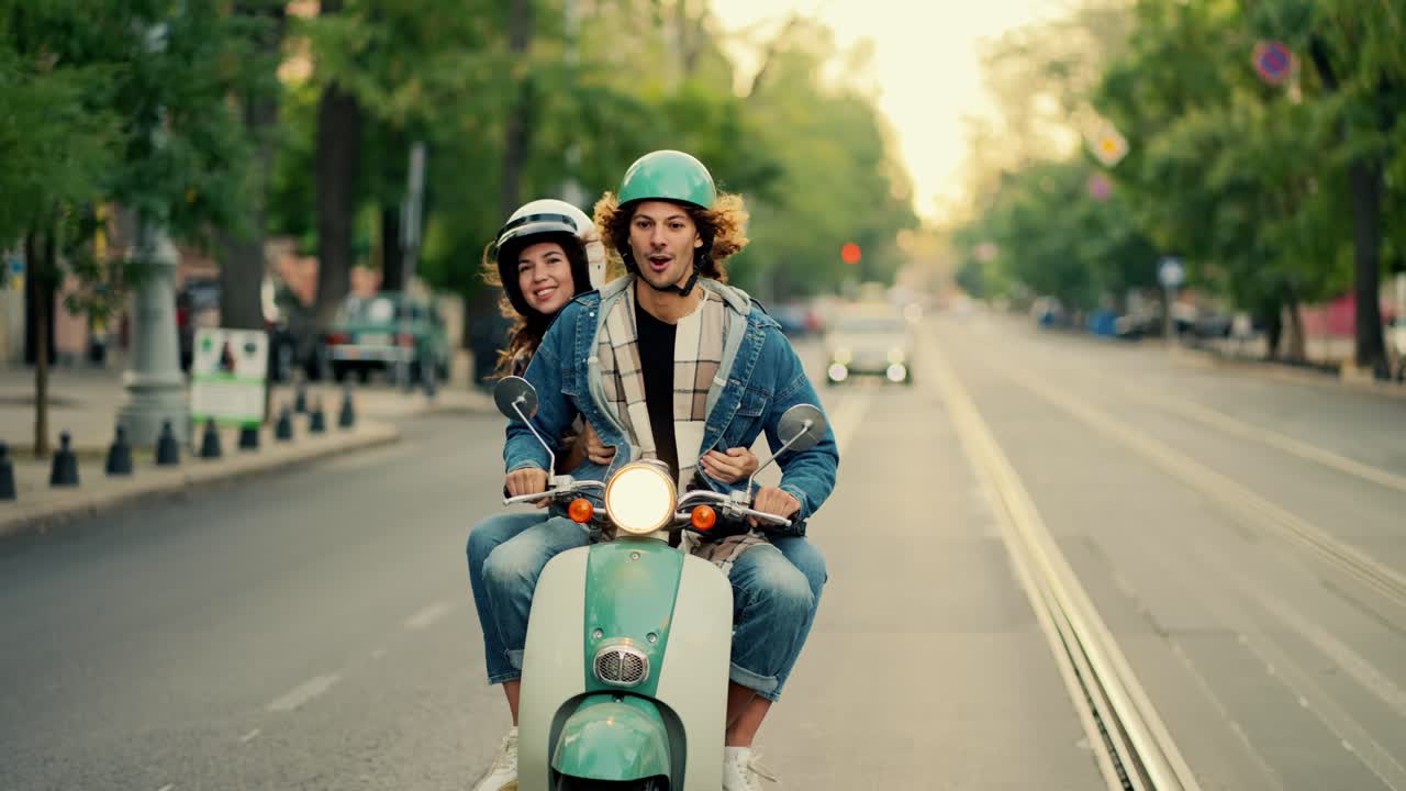 A happy guy with long curly hair in a denim jacket rides on his green moped with the headlights on, he is surprised by the bumps in front of him and from behind the guy’s shoulder his happy girlfriend in a white helmet for a moped looks out in the summer on a wide street in the city