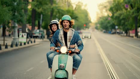 A-happy-guy-with-long-curly-hair-in-a-denim-jacket-rides-on-his-green-moped-with-the-headlights-on,-he-is-surprised-by-the-bumps-in-front-of-him-and-from-behind-the-guy’s-shoulder-his-happy-girlfriend-in-a-white-helmet-for-a-moped-looks-out-in-the-summer-on-a-wide-street-in-the-city