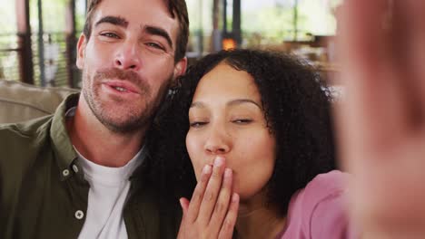 Mixed-race-couple-waving-and-blowing-kisses-on-video-call-sitting-on-the-couch-at-vacation-home