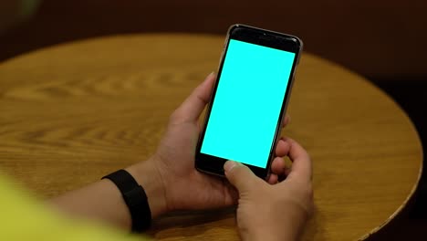 over shoulder view of one hand holding green screen smartphone on patterned wooden table.