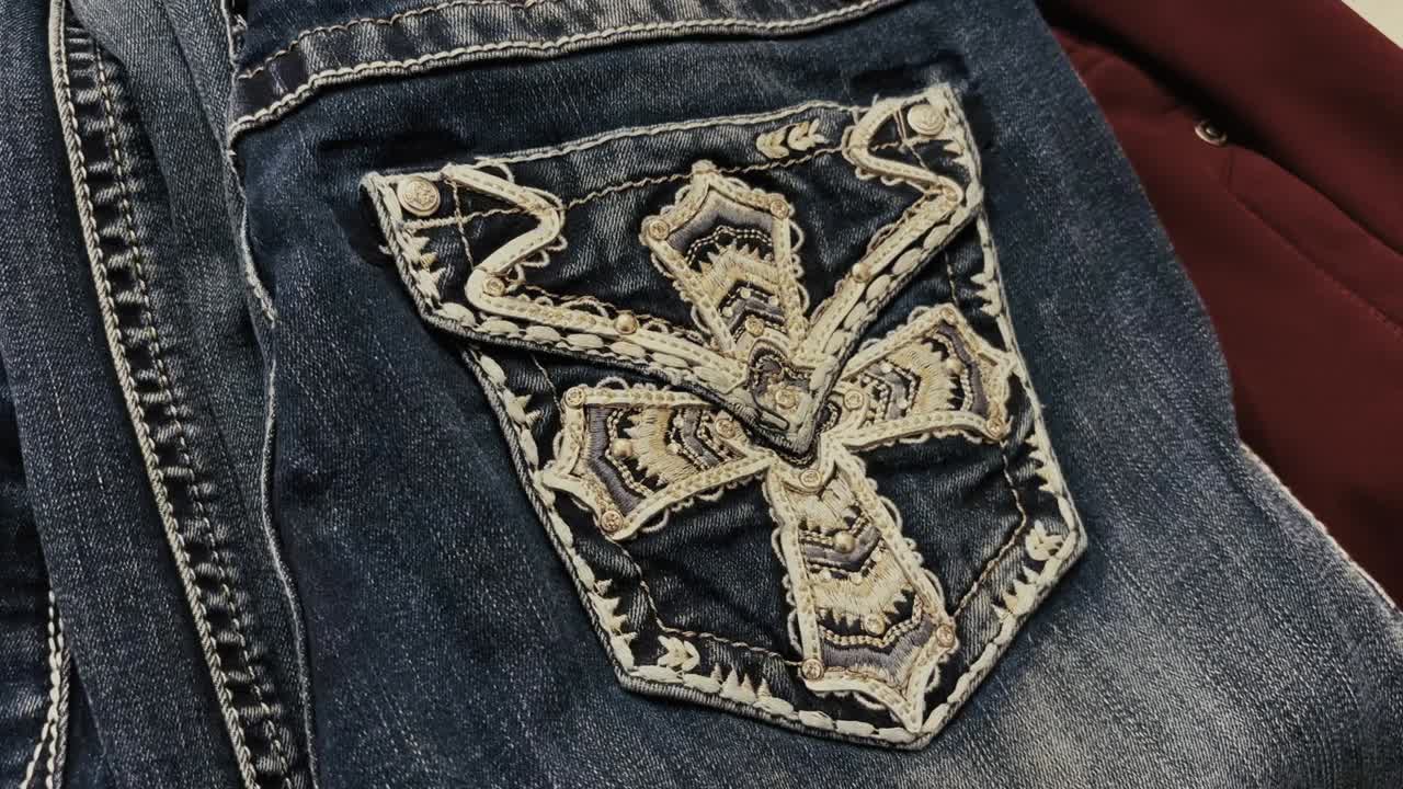 Detailed view of a denim pocket featuring an ornate stitched cross design with layered embroidery and textured thread patterns