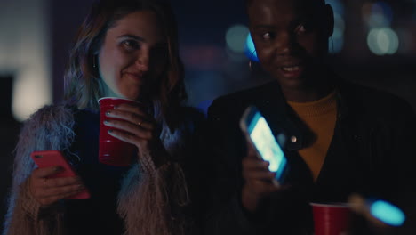 group of friends relaxing on rooftop at night using smartphone technology showing beautiful women sharing social media lifestyle enjoying weekend party