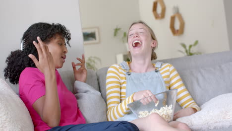 amigas adolescentes felices y diversas hablando y comiendo palomitas de maíz en el sofá en casa, en cámara lenta