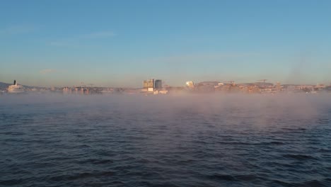 aerial low flying over waters with floating fog rising to reveal bjorvika in sentrum borough of oslo, norway