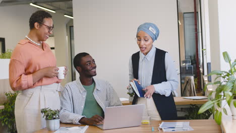 young worker working with laptop sitting at his desk while muslim businesswoman and businesswoman talk to him standing 1