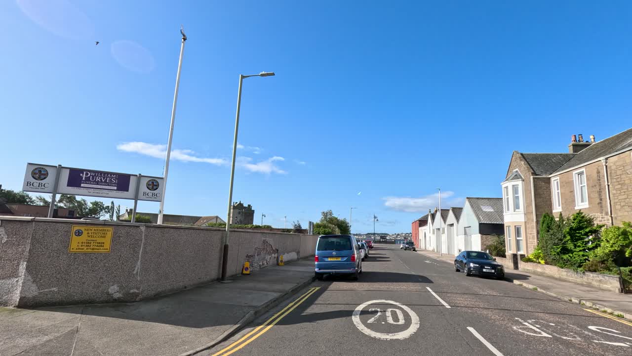 A vehicle travels along a quiet residential street lined with parked cars and stone buildings under clear blue skies in Broughty Ferry, Dundee