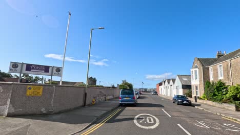 a vehicle travels along a quiet residential street lined with parked cars and stone buildings under clear blue skies in broughty ferry, dundee
