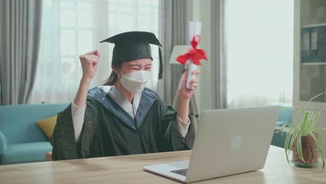 asian woman wearing protection face mask and celebrating with university certificate to the family during an online video call. pretty female graduate wearing a graduation gown and cap sitting on the living room