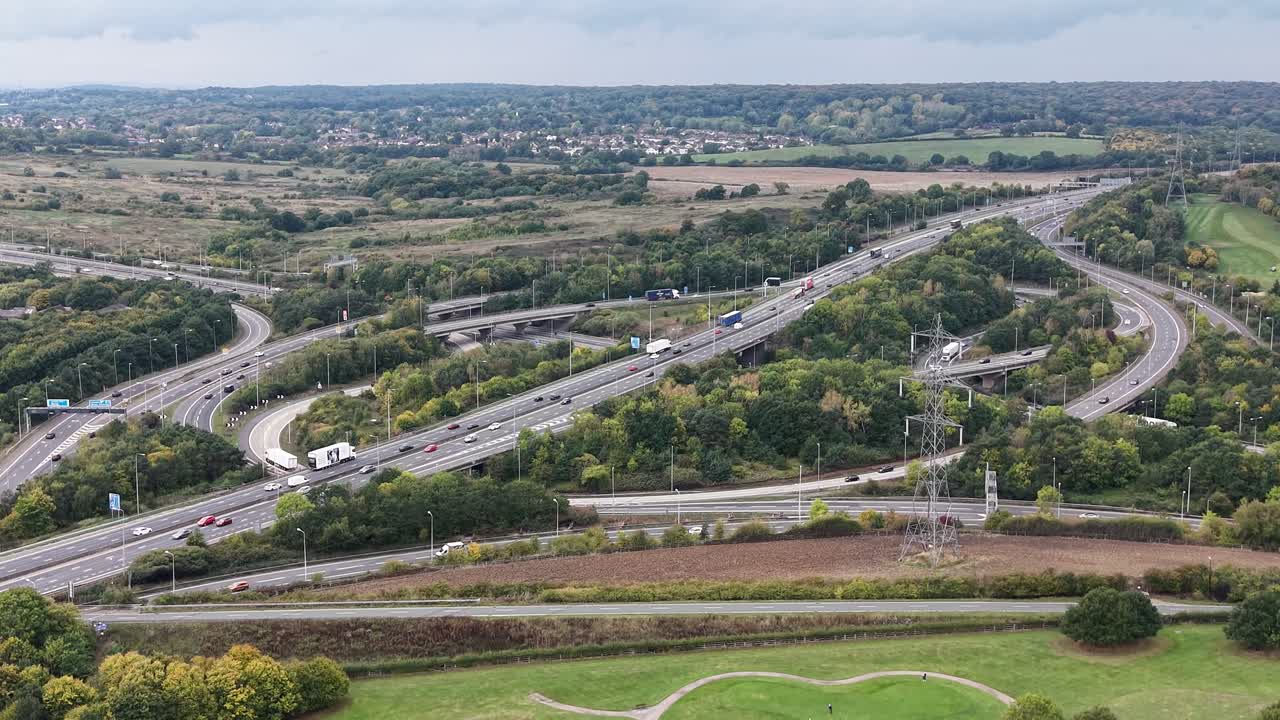 Aerial drone footage of the busy M25 and M11 motorway junction in the UK, showing heavy traffic movement and a golf course in the foreground. infrastructure surrounded by green countryside