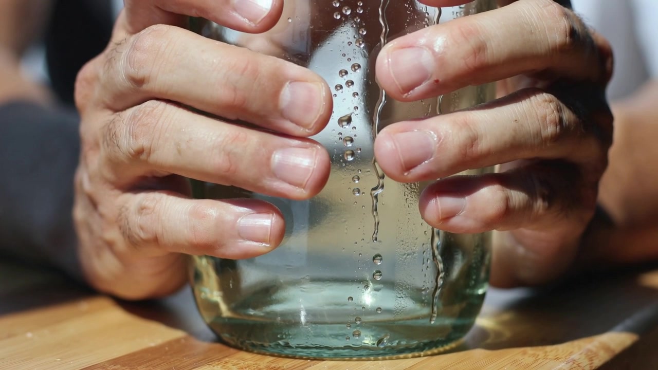 Mani che si chiudono attorno a un barattolo di vetro su un tagliere di legno per stabilizzarlo, facendo scivolare goccioline d'acqua verso il basso.