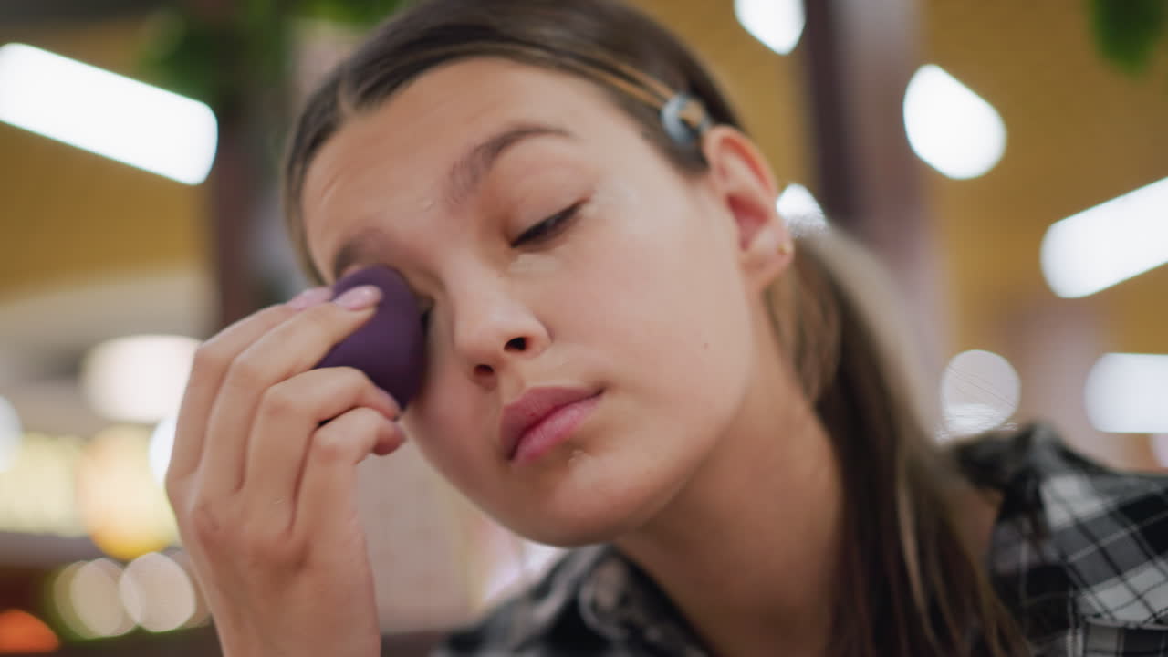 primer plano de una mujer joven mezclando la base debajo de los ojos usando esponja de belleza, expresión enfocada, piel lisa y aplicación de maquillaje precisa, destacando la rutina de belleza y las técnicas cosméticas
