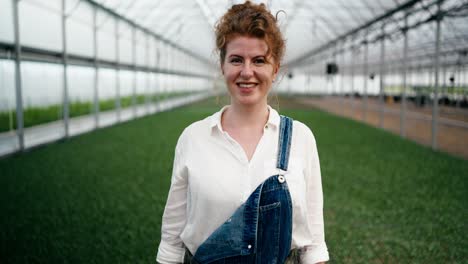 portrait of a happy woman with red curly hair in a white shirt who works on a farm among plants with a watering can in her hands and poses