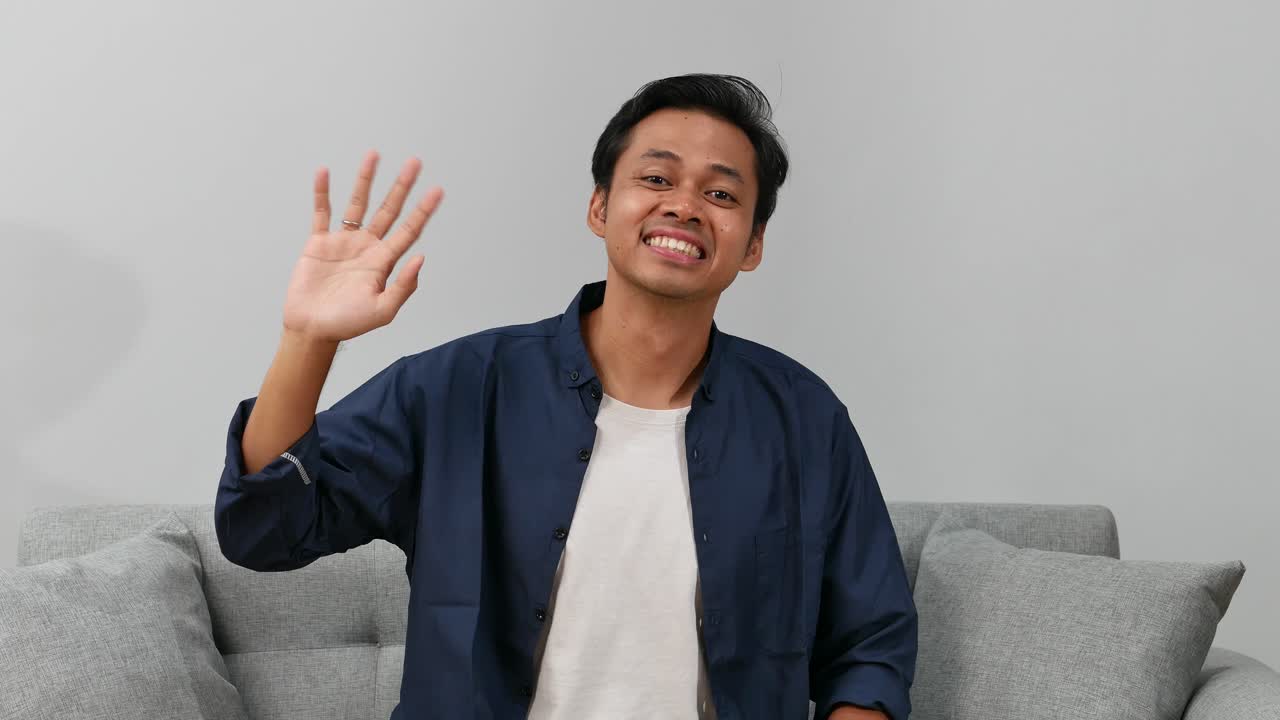 Happy friendly young man waves palm in hello gesture welcomes someone with hospitable expression at home